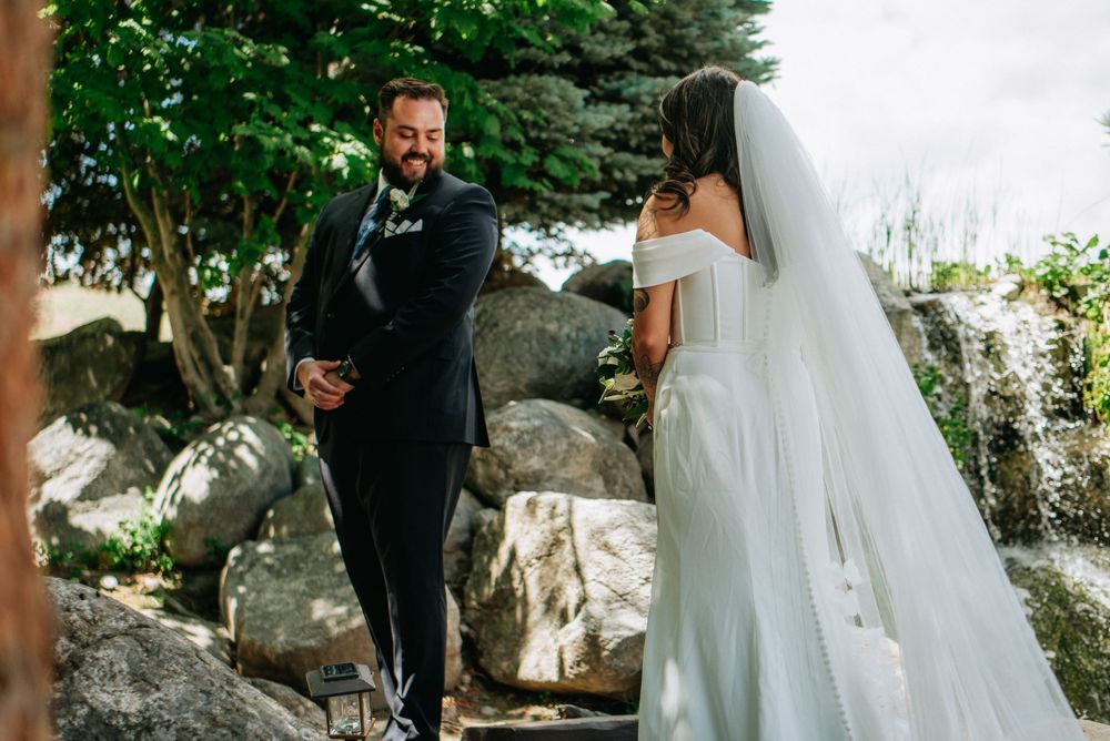Groom in navy suit smiling at bride in white off-shoulder gown and veil during outdoor wedding ceremony.