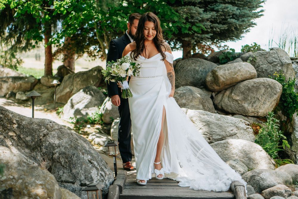 Bride in white gown with slit holds bouquet beside groom on rocky garden steps outdoors.