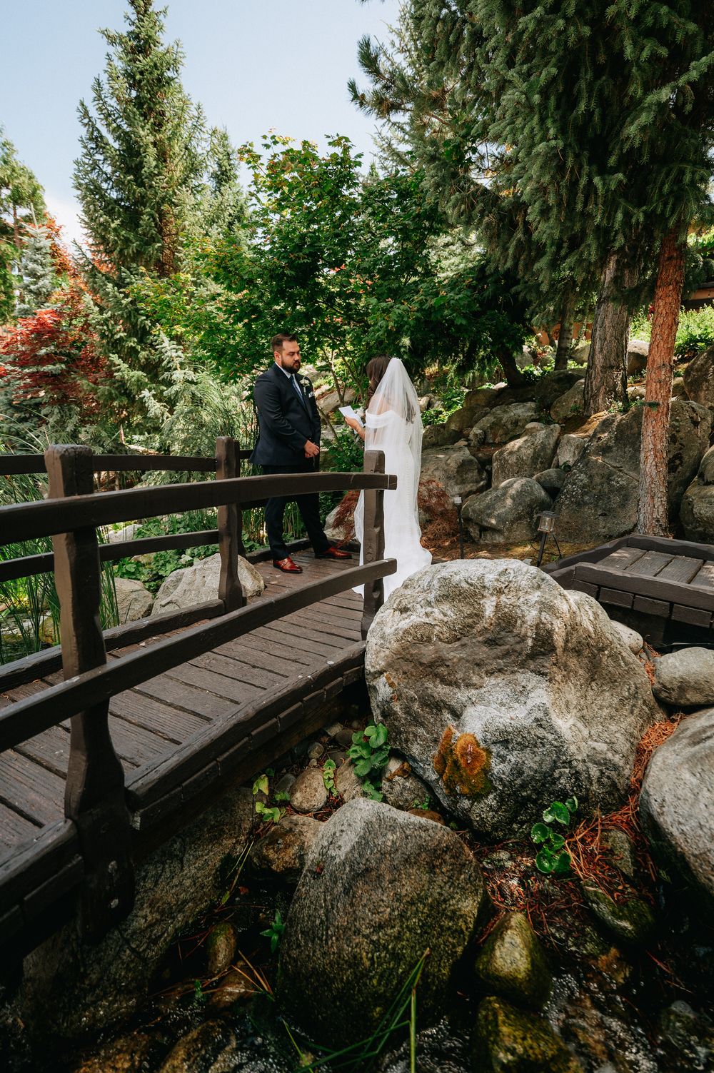 Bride and groom share vows on a wooden bridge surrounded by lush greenery and rocks in a garden wedding setting.