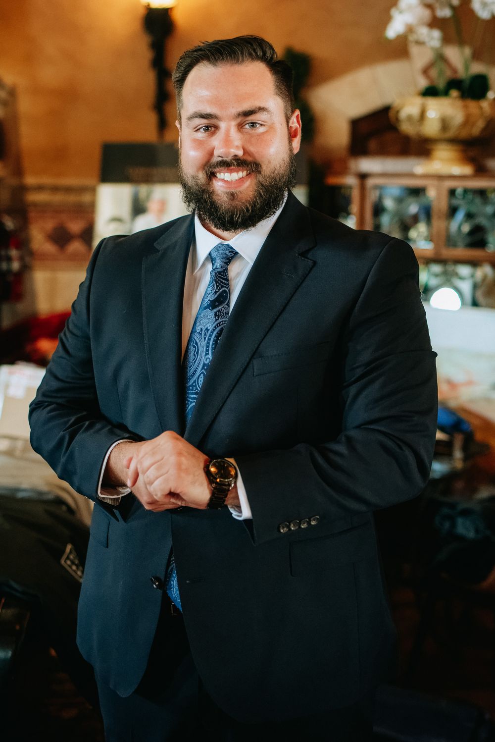 Smiling bearded man in dark suit and blue tie adjusting cufflinks in an elegant restaurant interior.