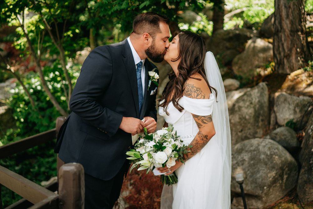 Bride and groom share a kiss outdoors, bride holding white bouquet, groom in navy suit, rocky garden backdrop.