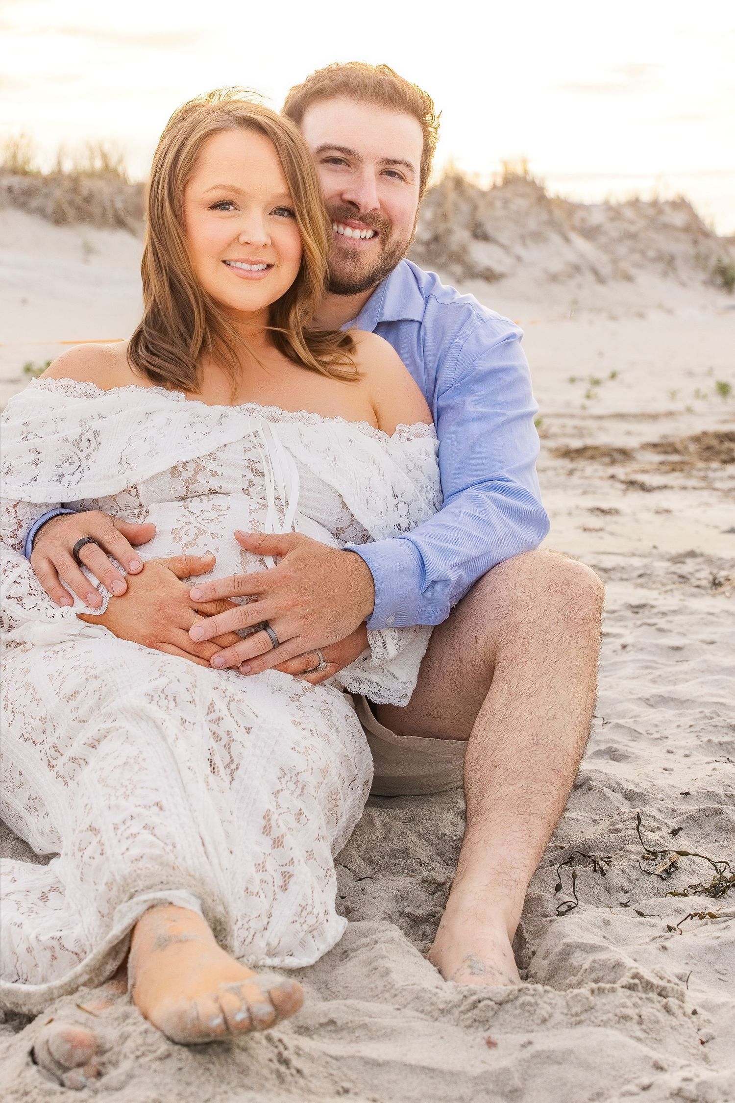 Pregnant couple sitting on sandy beach, woman in white lace dress, man in blue shirt, both smiling and cradling baby bump.