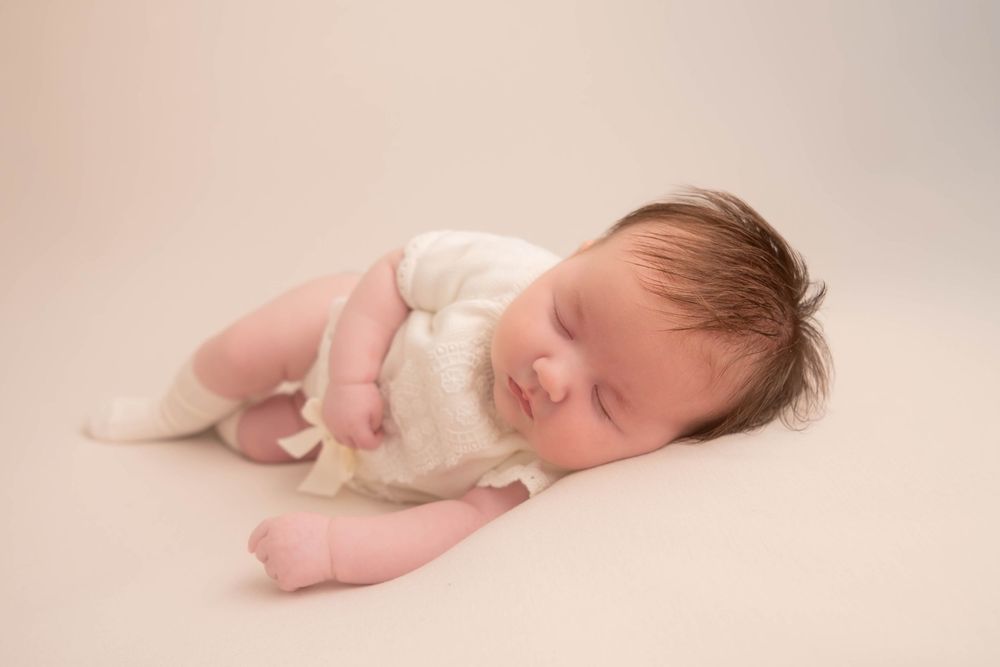 Peaceful newborn baby sleeping on cream backdrop wearing white romper, showcasing delicate features and dark hair.
