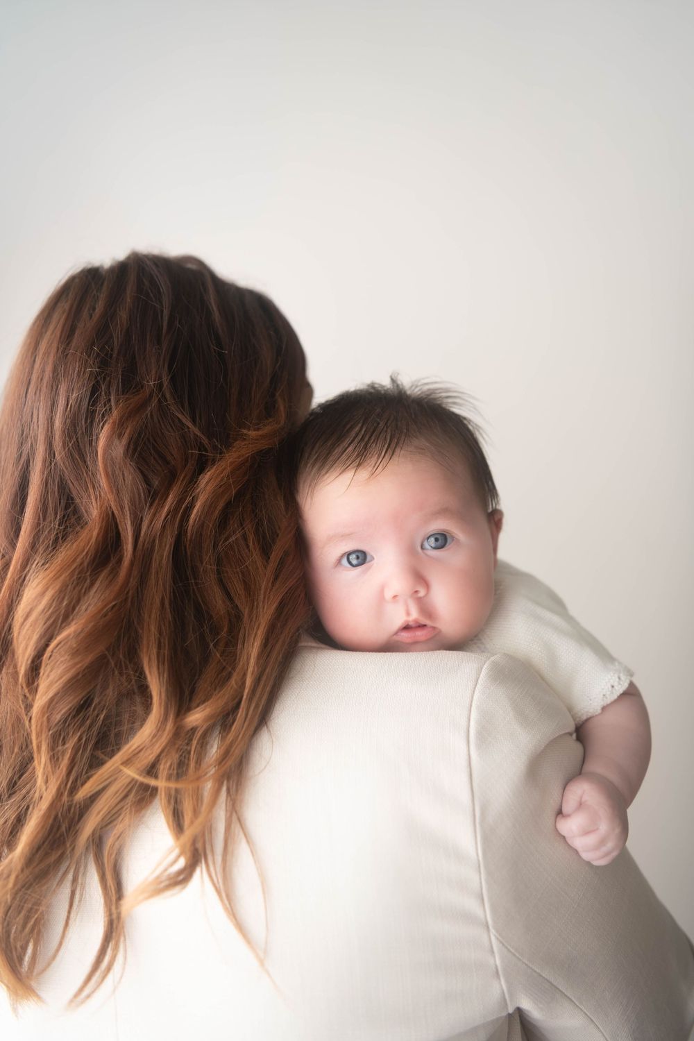 A mother with long auburn hair holds a blue-eyed newborn baby in a white onesie against her shoulder.