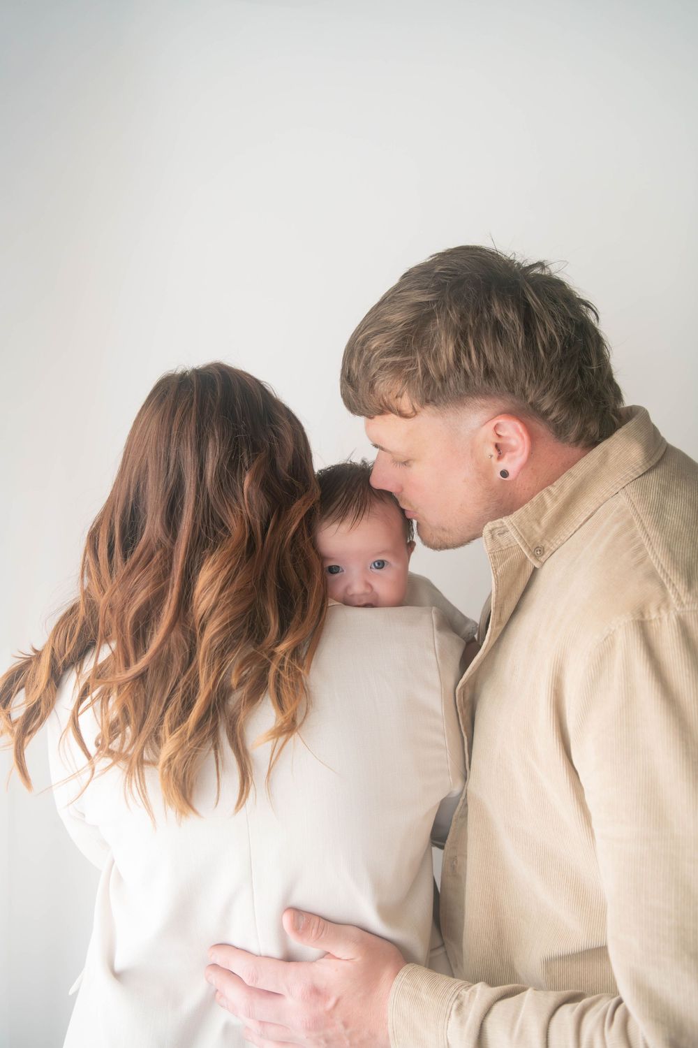A couple in neutral tones tenderly kisses their newborn baby wrapped in a white swaddle against a light background.