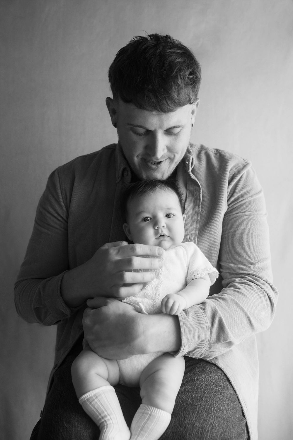 Black and white portrait of a young father tenderly holding his newborn baby, both looking down in a studio setting.