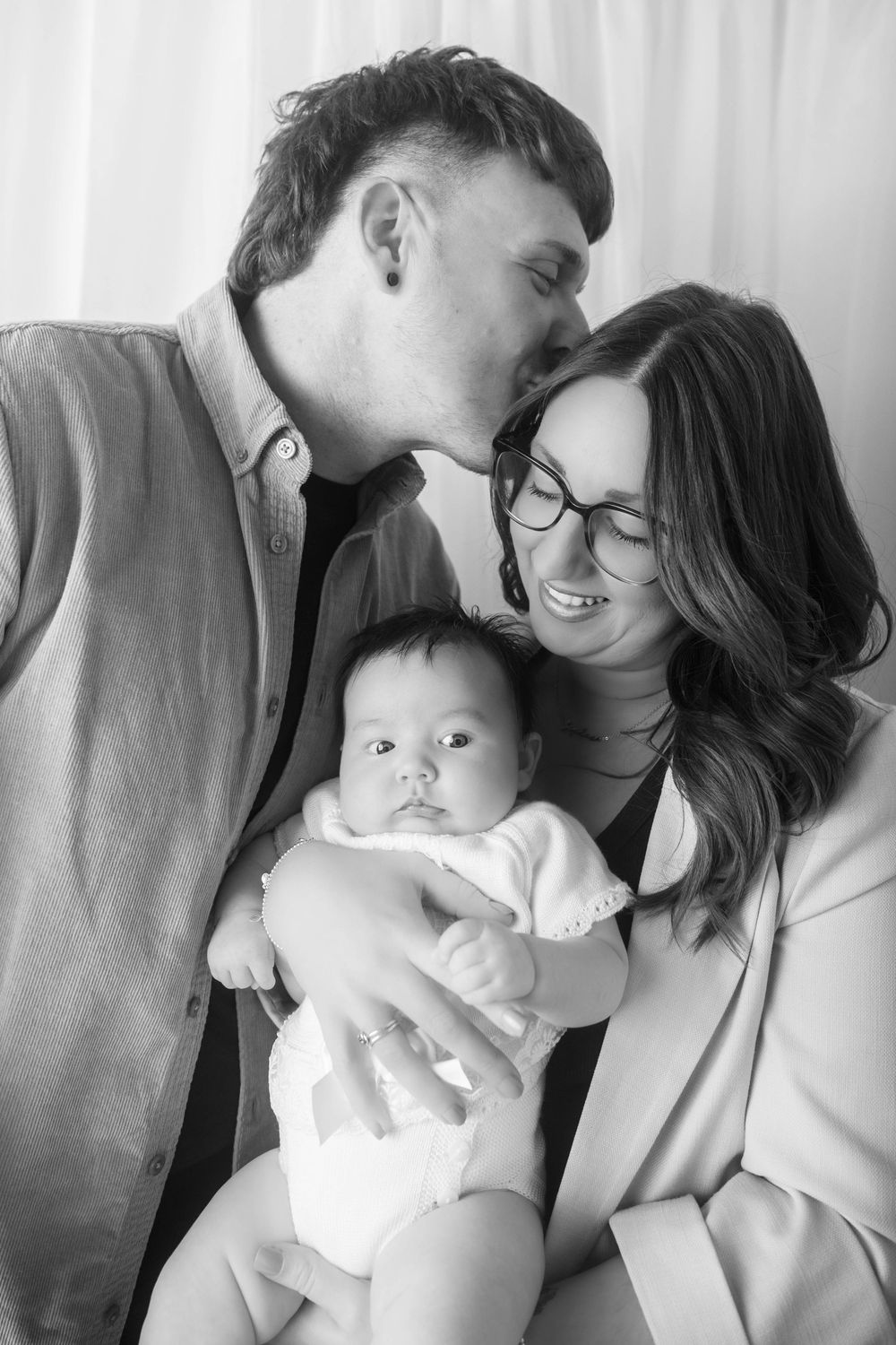 Black and white photo of a couple kissing and smiling while holding their newborn baby in a studio for a family photography session.