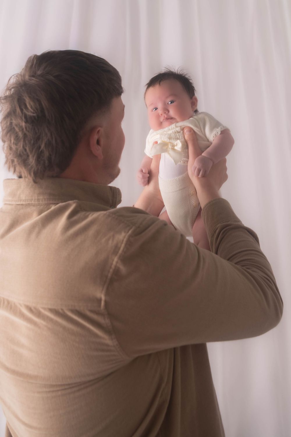 A father holds his newborn baby up in the air, gazing lovingly at the infant against a soft white backdrop.