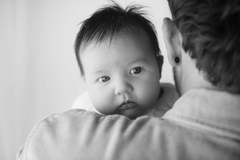 Black and white photo of a baby resting on an adult's shoulder, looking at the camera with curious eyes. family photography Caerphilly
