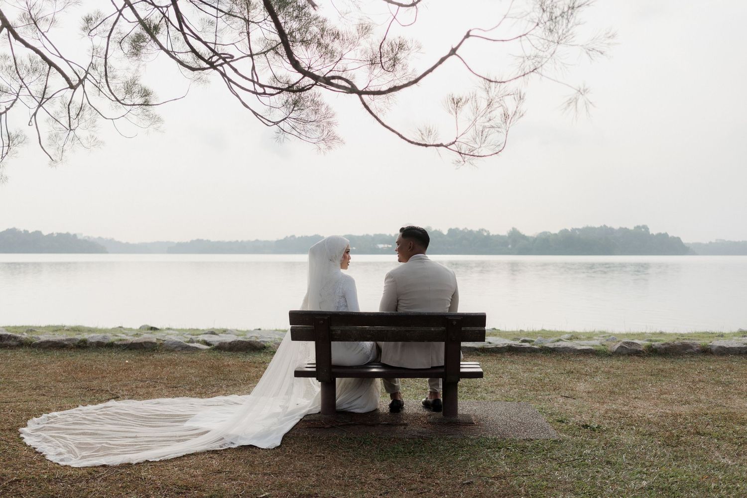 Newlywed couple in wedding attire sitting on a bench overlooking a serene lake surrounded by trees.