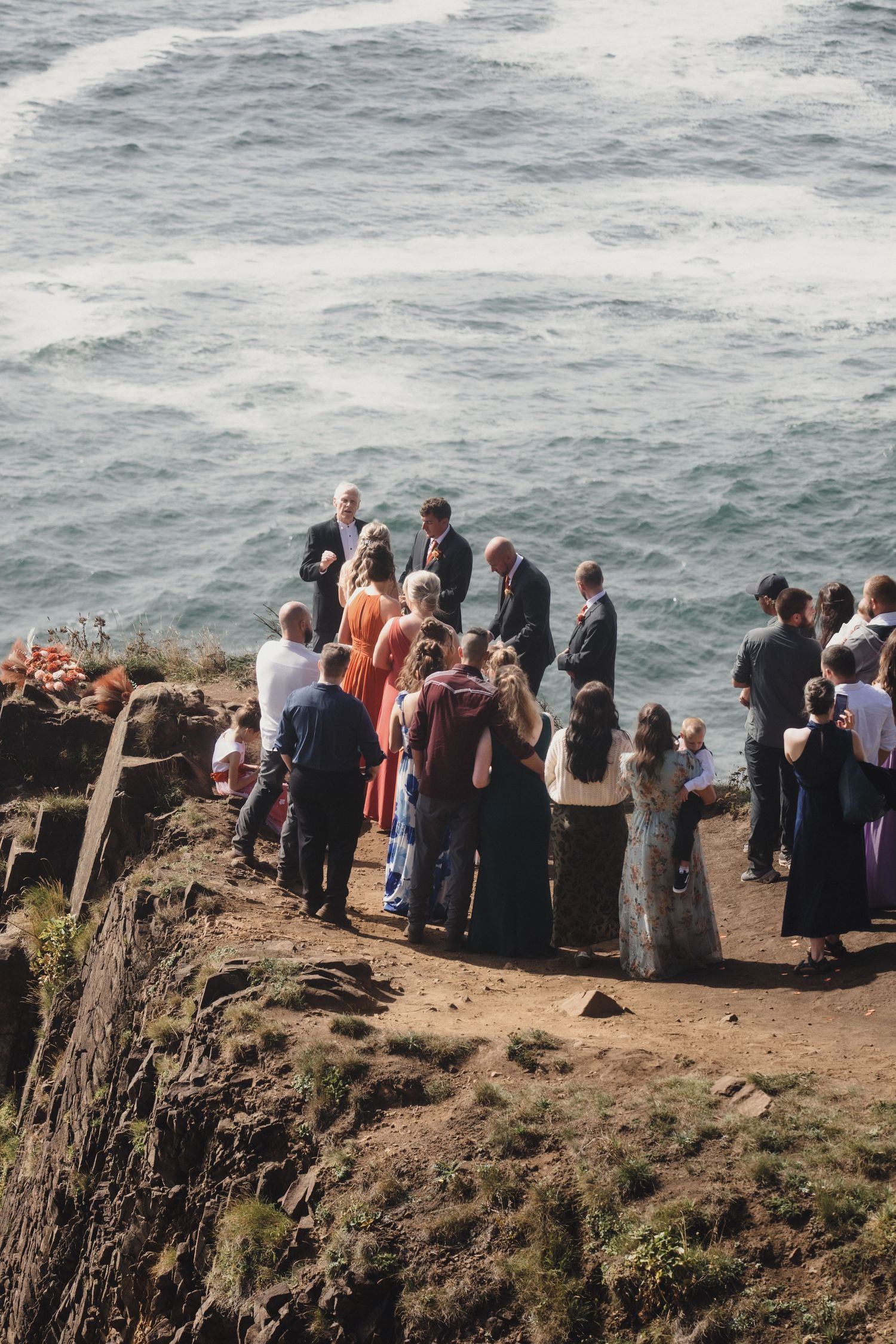 Small wedding ceremony group gathered on a rocky coastal cliff overlooking dramatic ocean waves below.