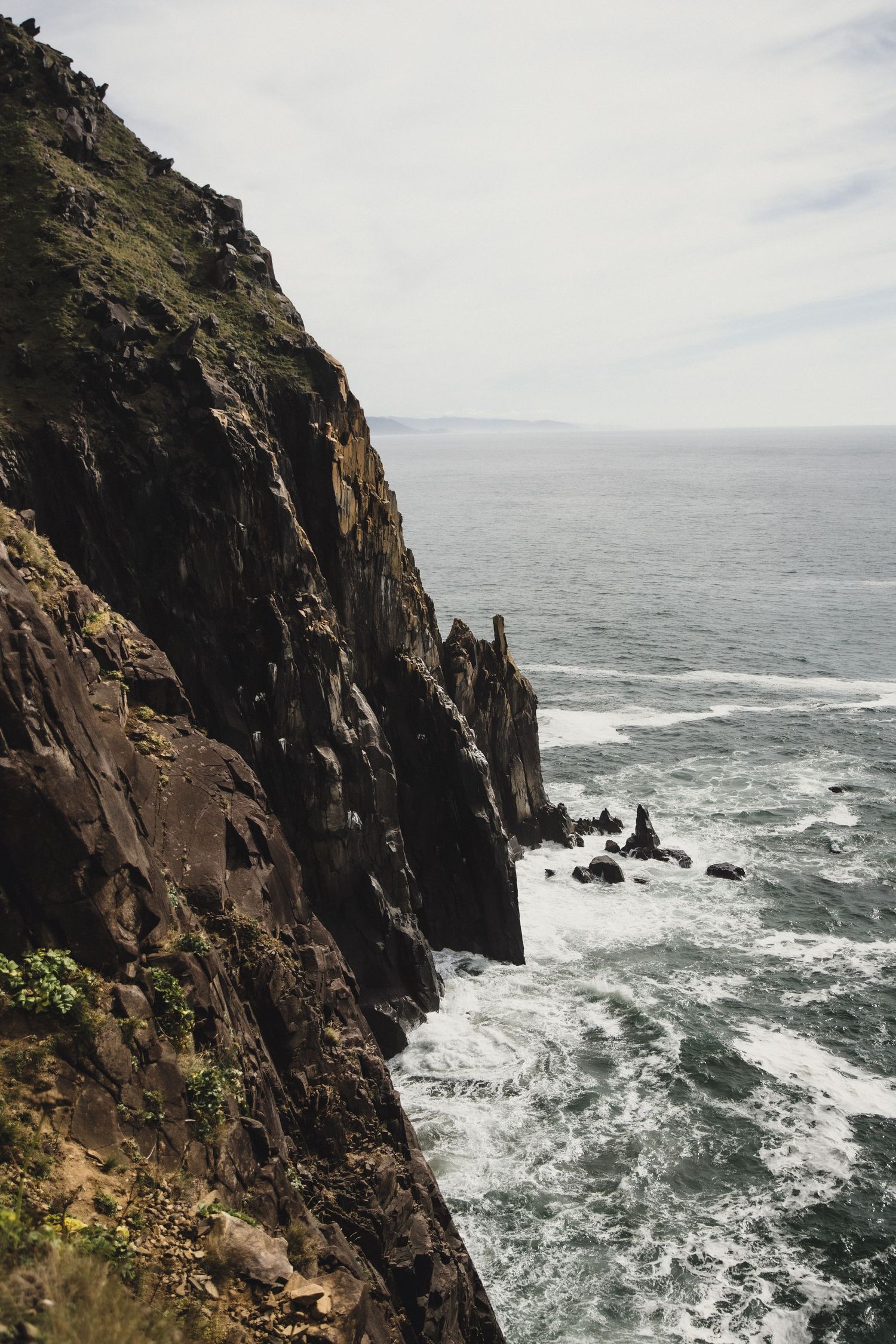 Dramatic coastal cliffs with dark rocky faces drop into churning ocean waves under a cloudy sky.