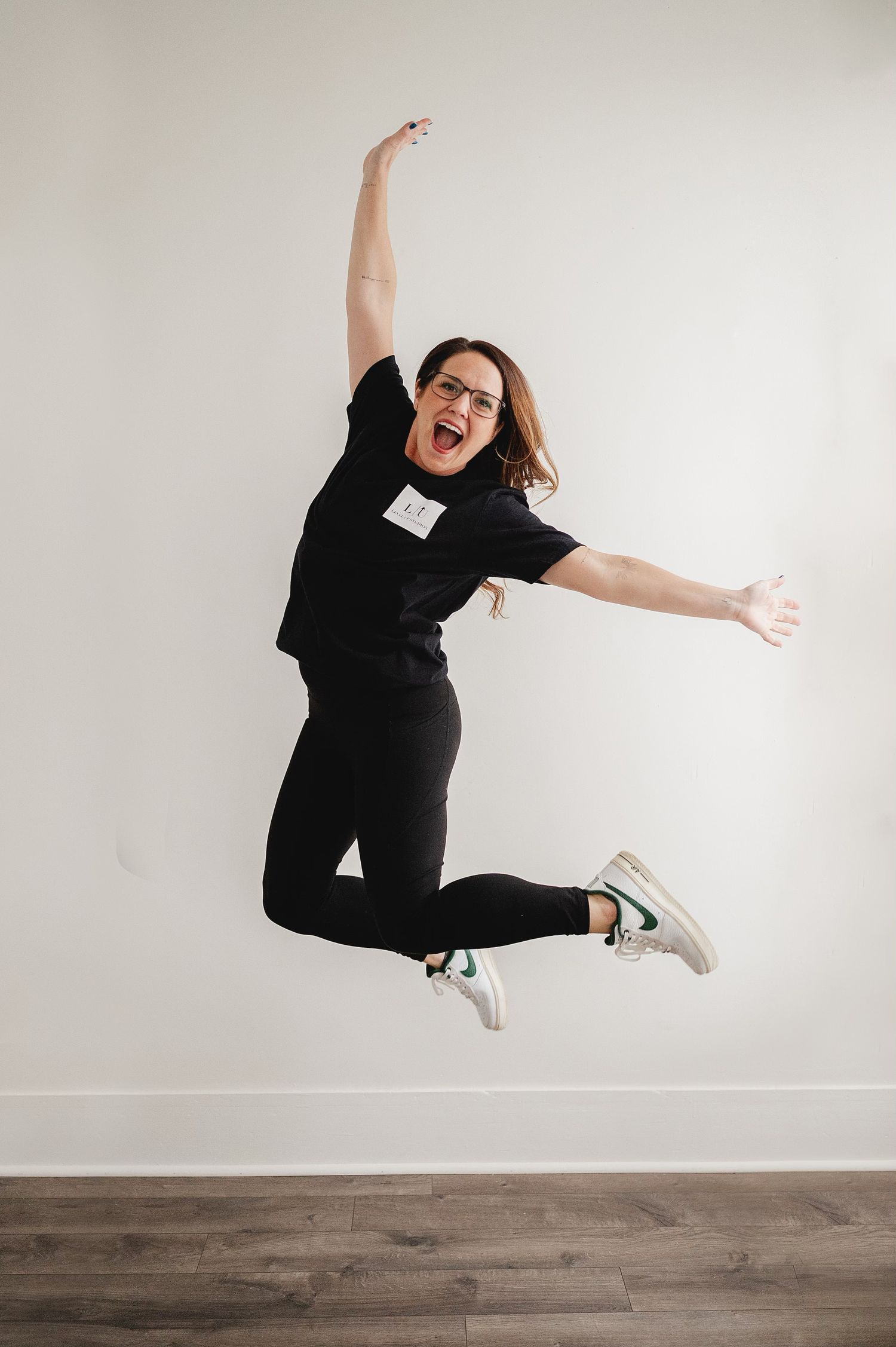 Woman in black outfit jumping joyfully with arms raised, wearing glasses and green sneakers against white wall.