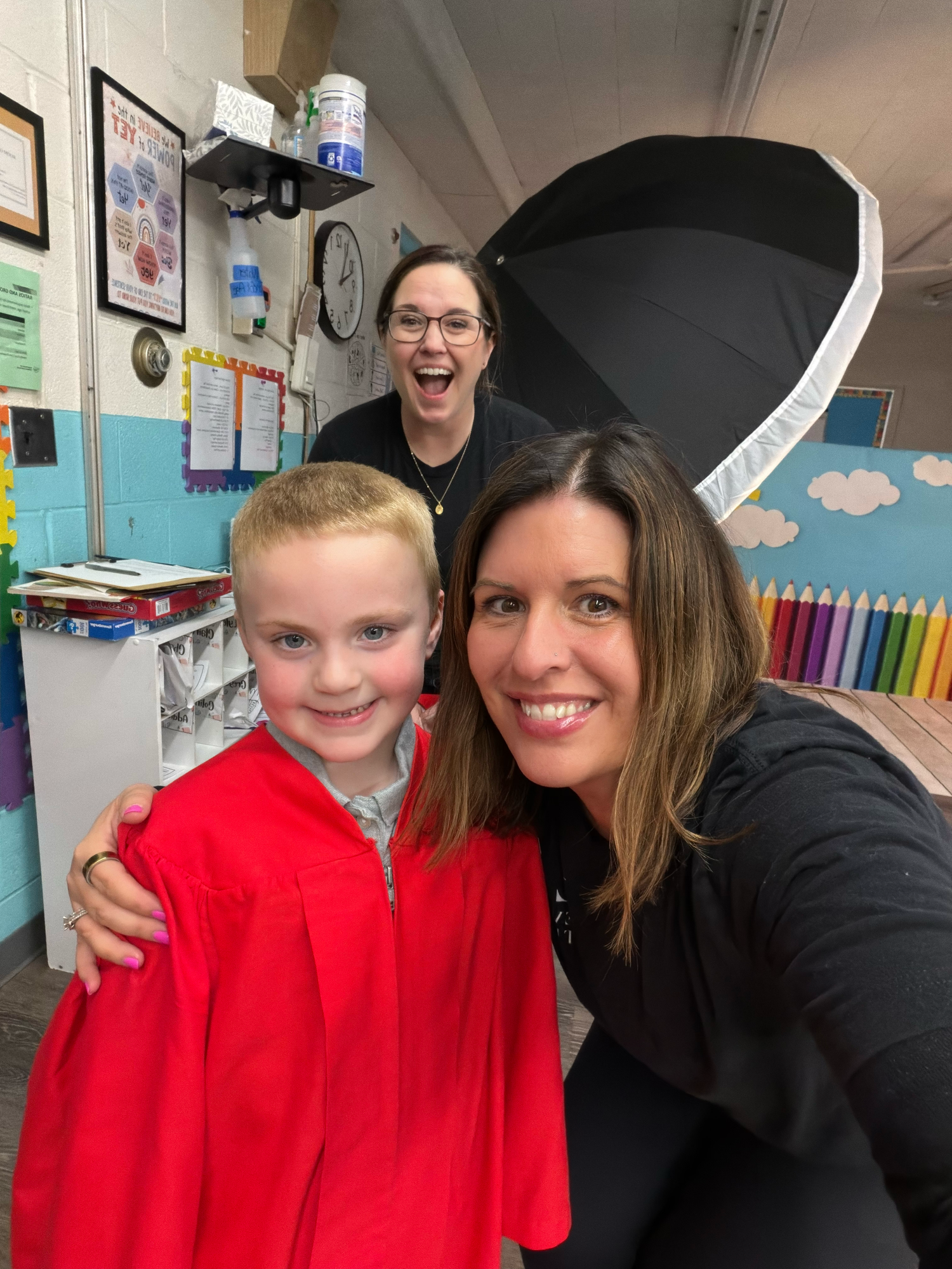 Woman takes selfie with young child in red shirt and laughing teacher in colorful elementary classroom.