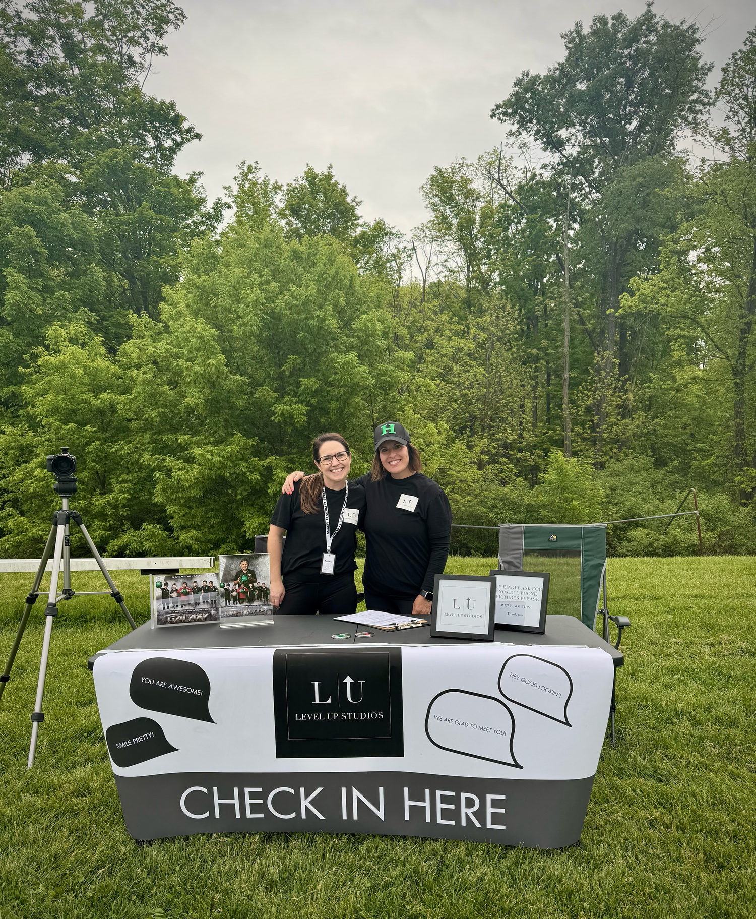 Two staff members stand behind a Level Up Louisiana check-in booth at an outdoor event surrounded by lush green trees.