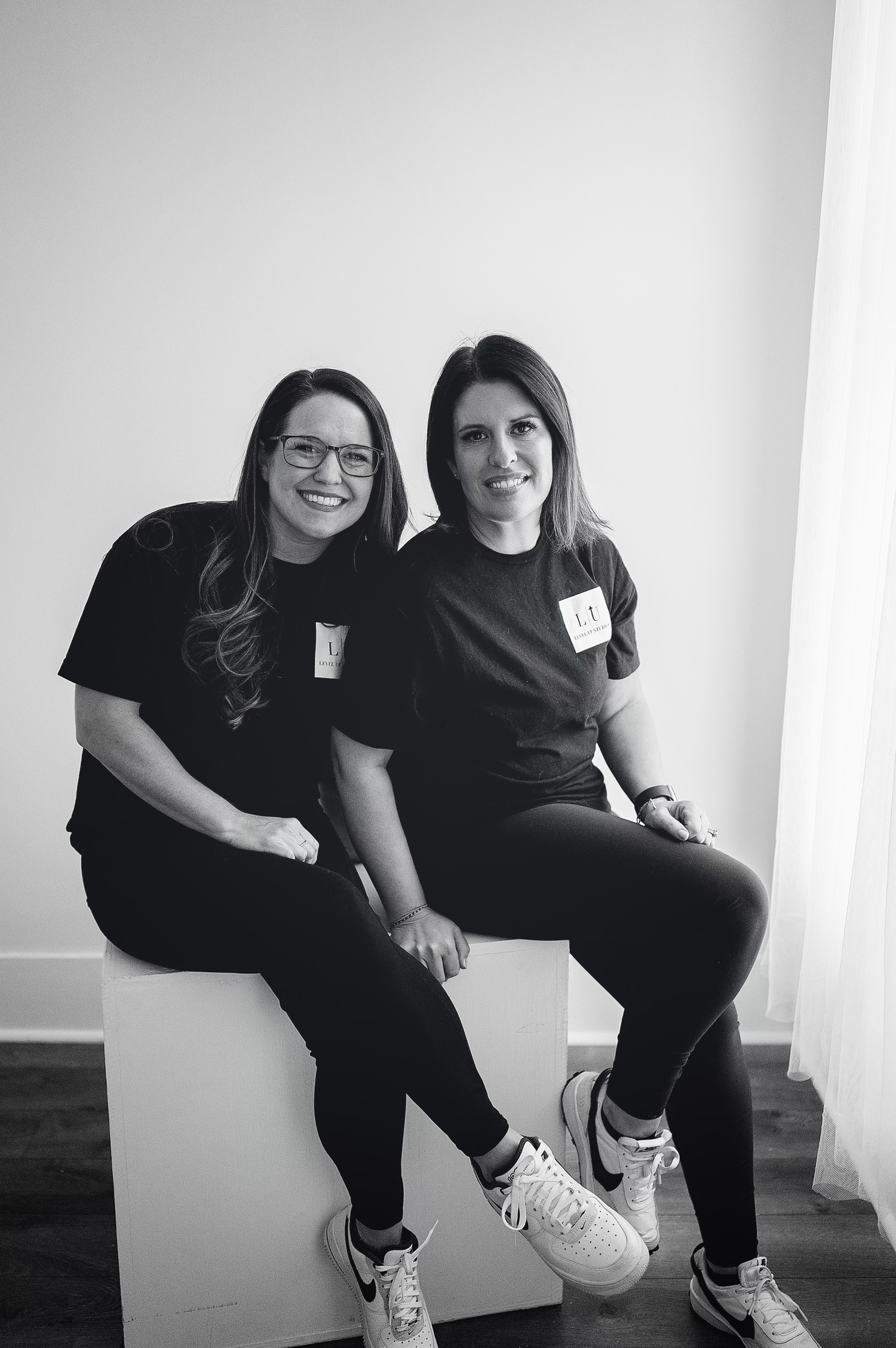 Two women in matching black outfits sit together smiling in a bright studio, black and white photo.