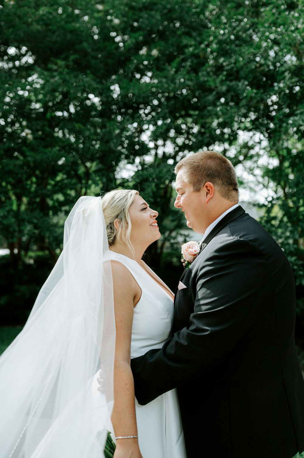Bride in white gown and veil gazes lovingly at groom in black suit outdoors among green trees.