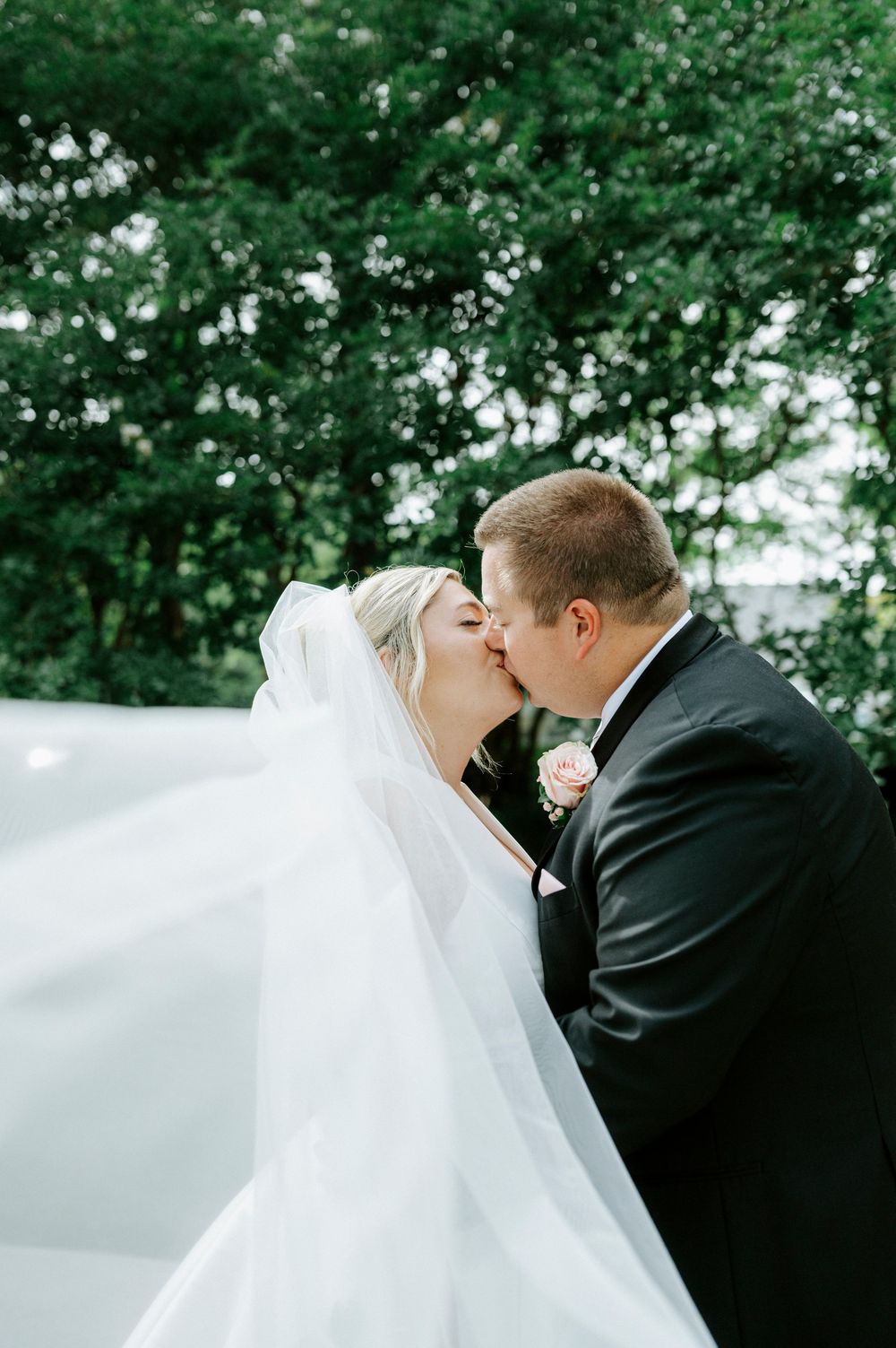 Bride and groom share a kiss as wedding veil flows dramatically in the wind outdoors.