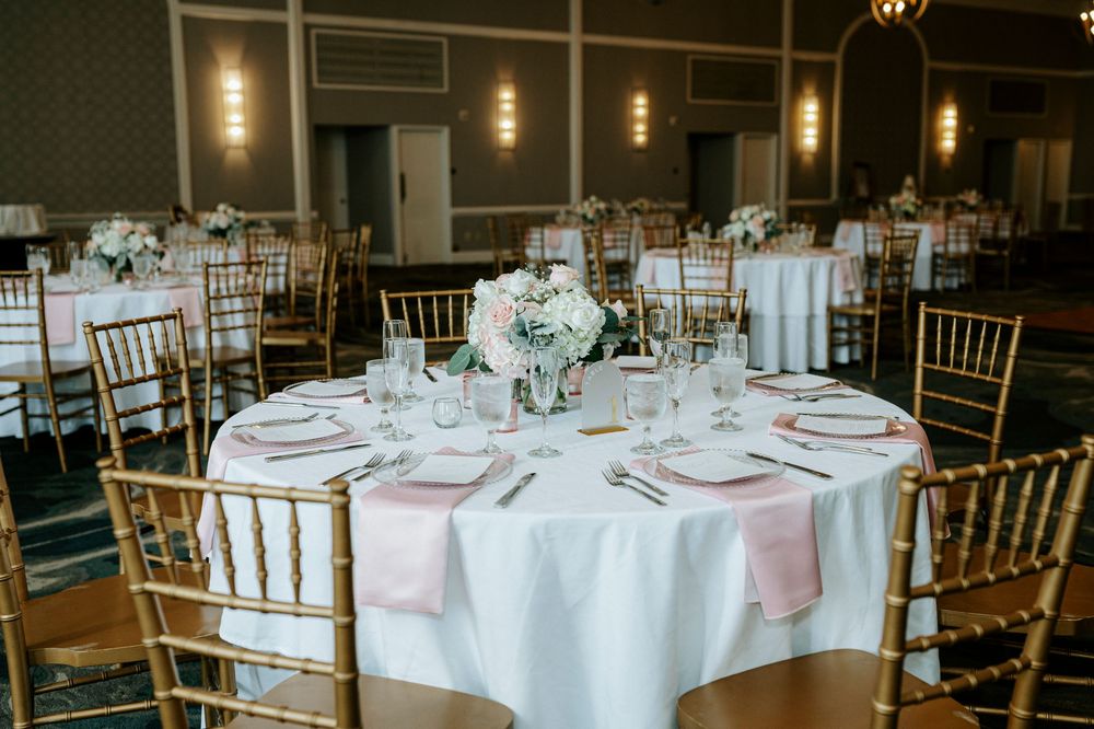 Wedding reception table with white linens, pink napkins, gold chiavari chairs, and floral centerpiece.