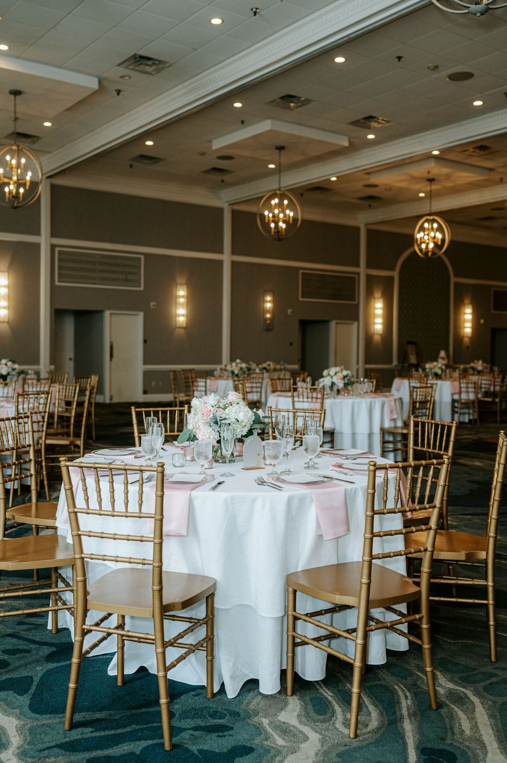 Elegant wedding reception ballroom with gold chiavari chairs, white tables, and globe chandelier lighting.