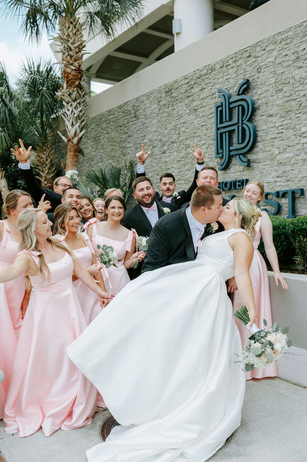 Bride and groom share a romantic dip kiss surrounded by cheering wedding party in pink bridesmaid dresses outside Hotel Ballast.