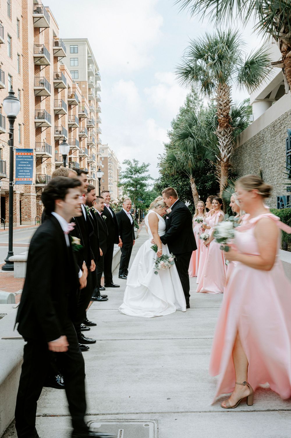 Wedding party lines up on urban sidewalk with palm trees as bride and groom share a tender moment in white and black attire.