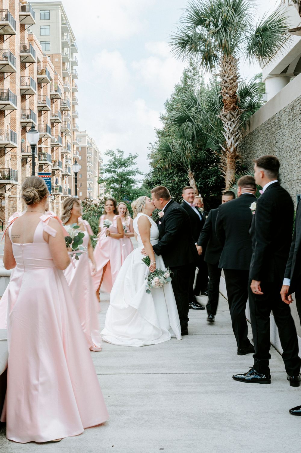 Wedding party in pink bridesmaid dresses and black tuxedos gathered outdoors near palm trees.