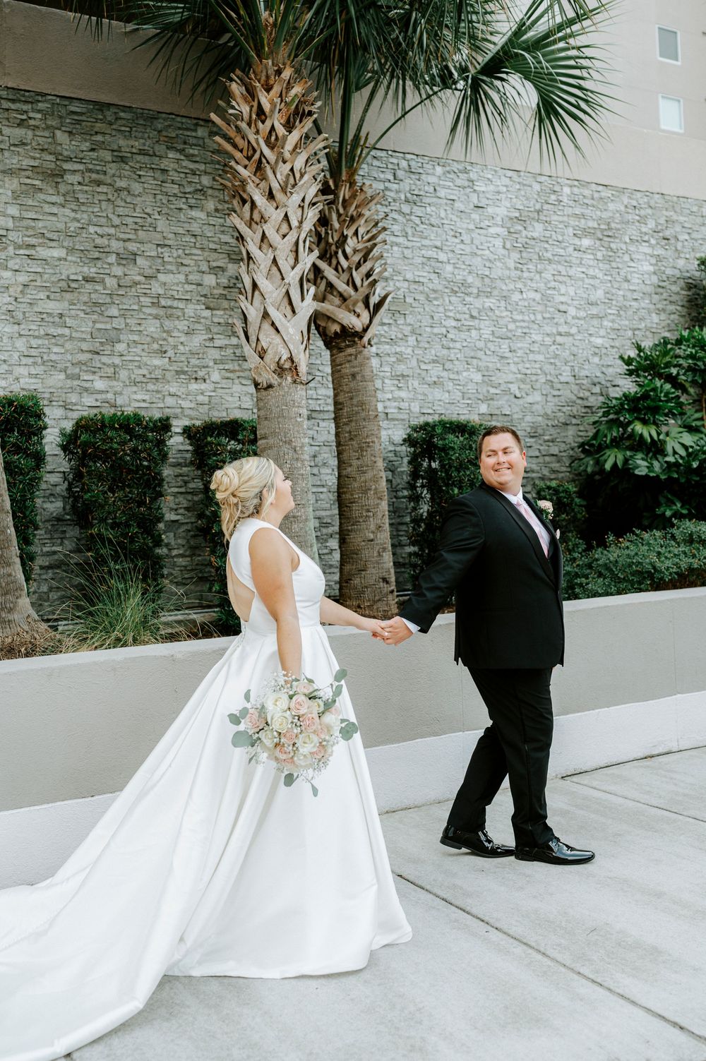 Bride in a white ball gown holds bouquet as groom in black tux leads her by the hand near palm trees.