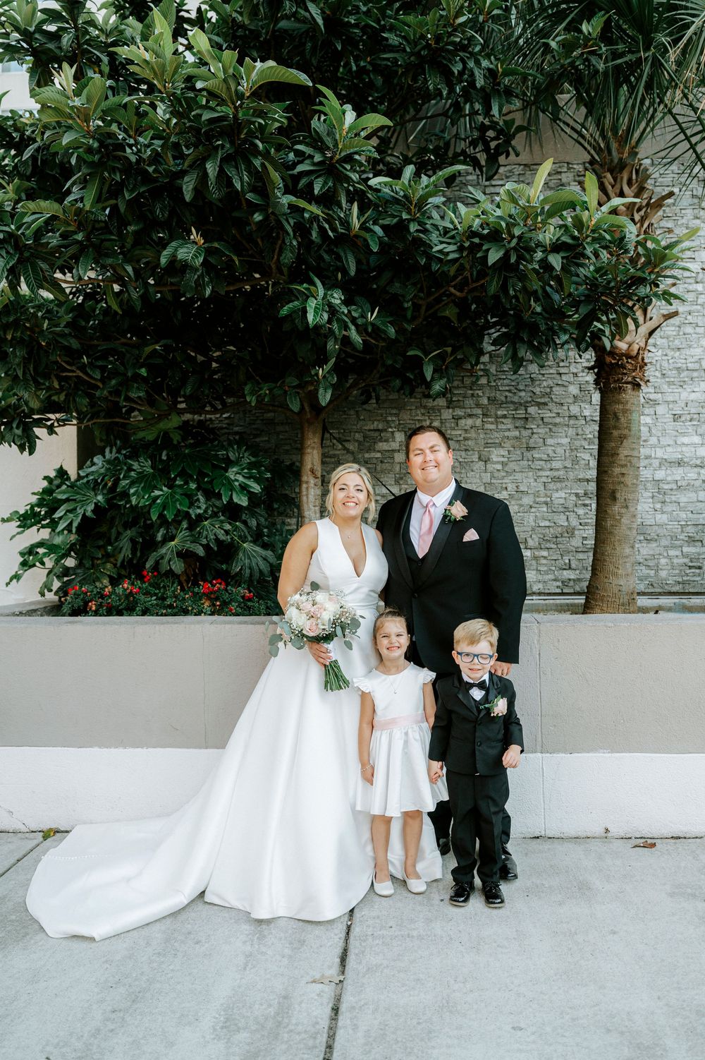 Bride and groom pose with flower girl and ring bearer outside amid lush green trees.