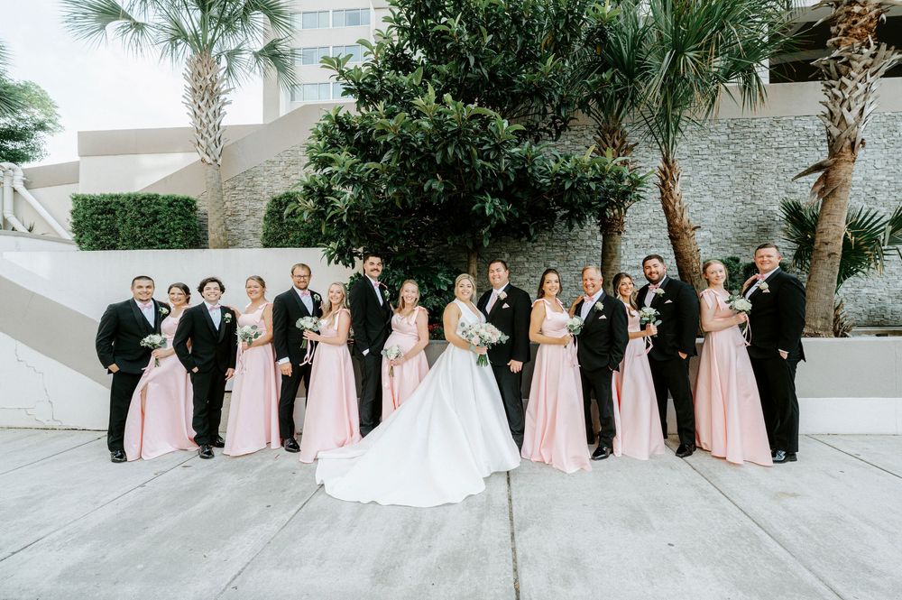 Wedding party poses outdoors in pink bridesmaid gowns and black tuxedos with bride and groom.