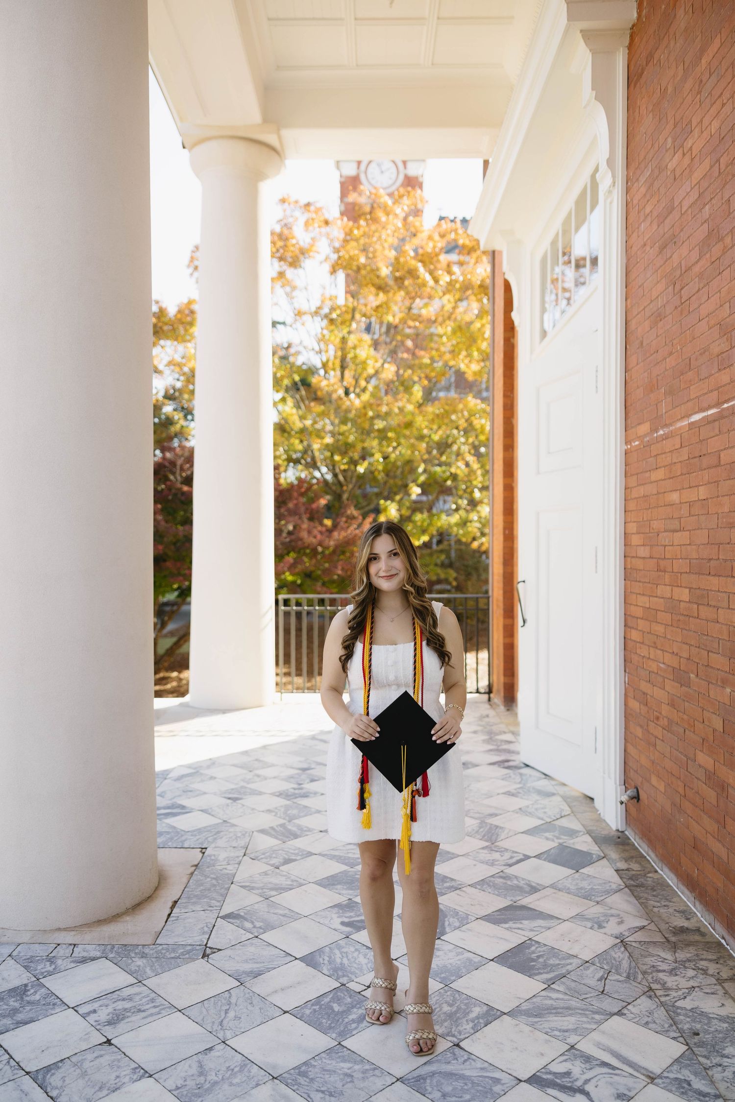 Young woman in white dress holding graduation cap with gold tassel, standing between white columns on a sunny campus.