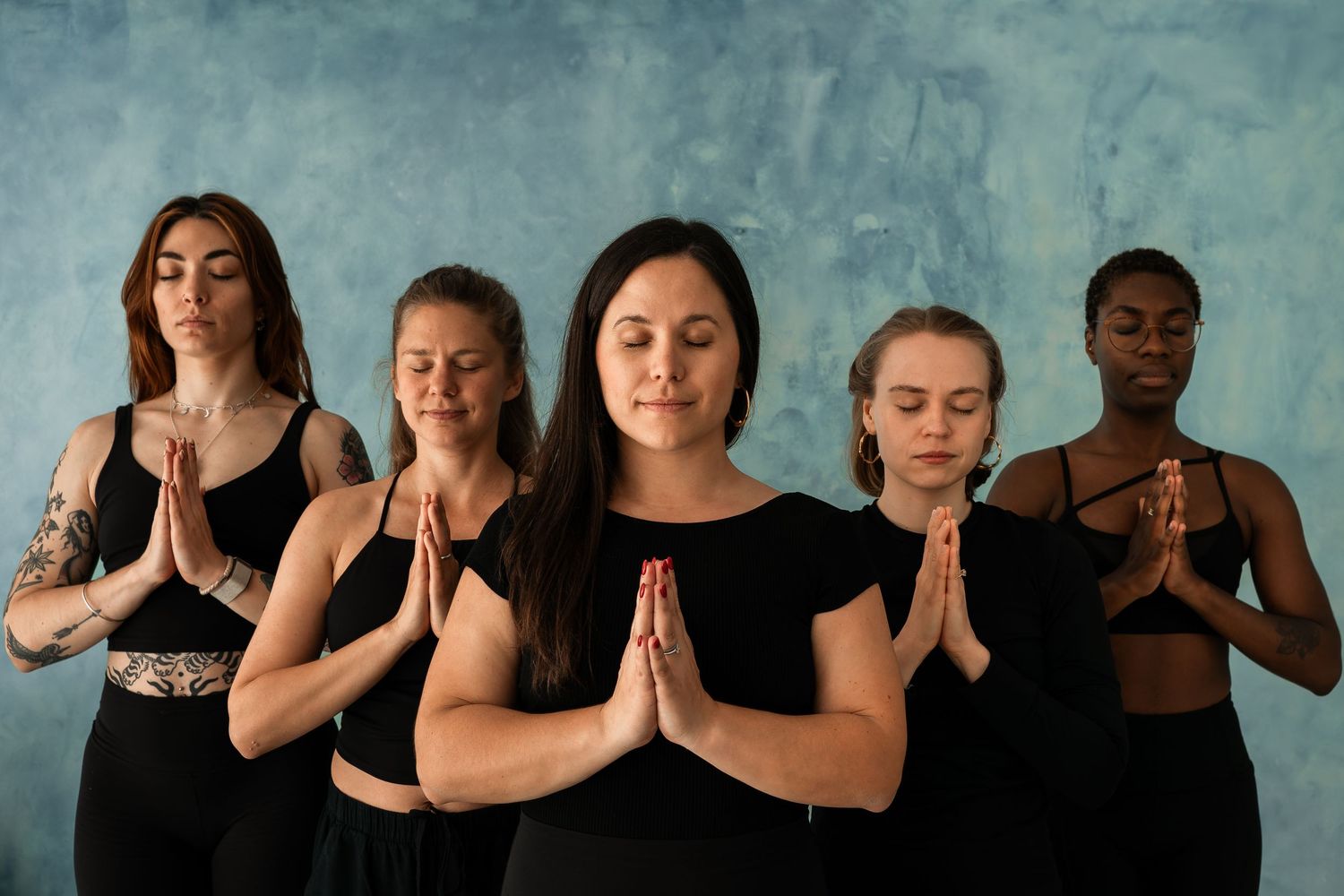 Five women in black activewear practice meditation in prayer pose with eyes closed against a blue wall.
