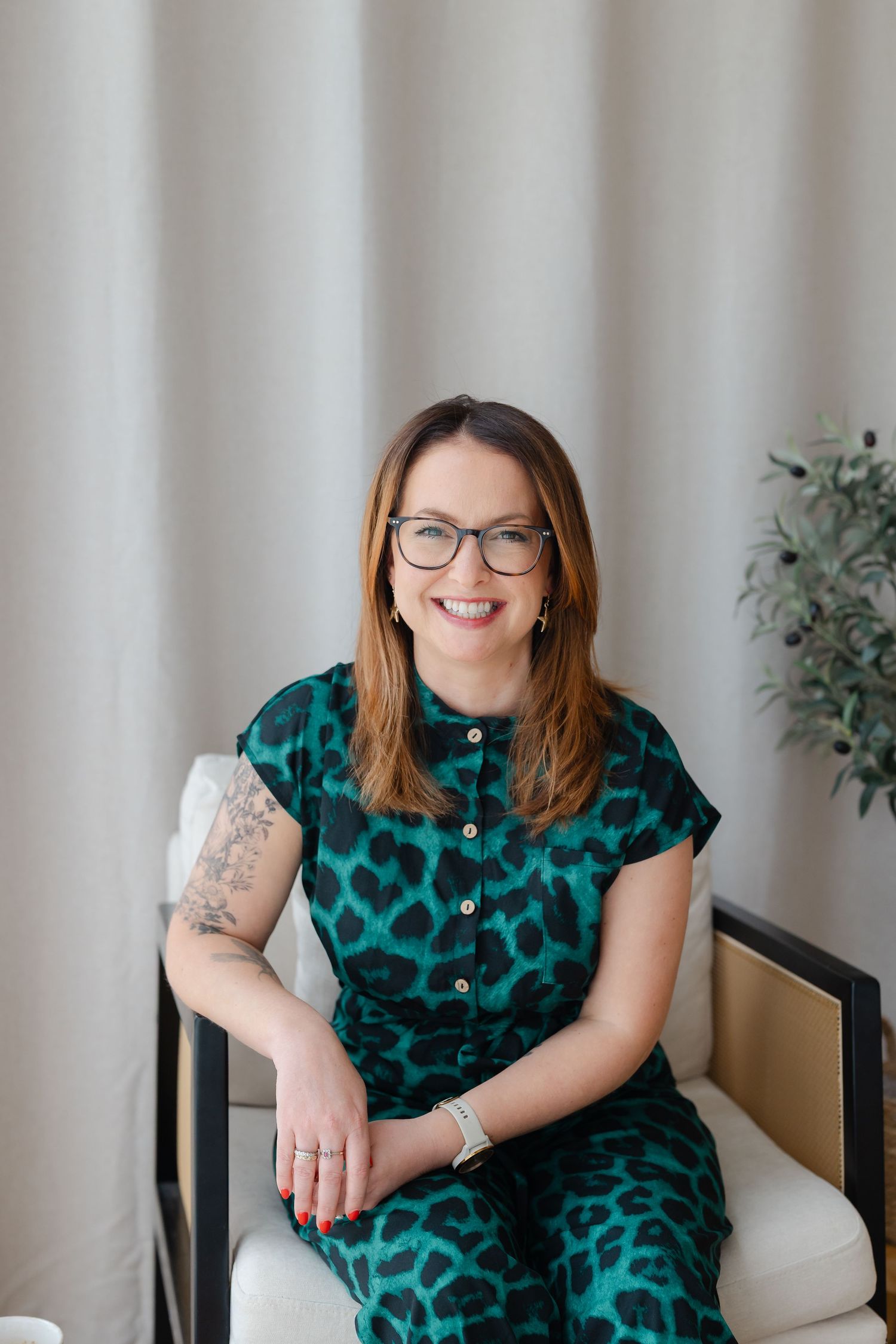 Woman in green leopard print jumpsuit sitting in chair, smiling, wearing glasses with tattoo visible on arm.