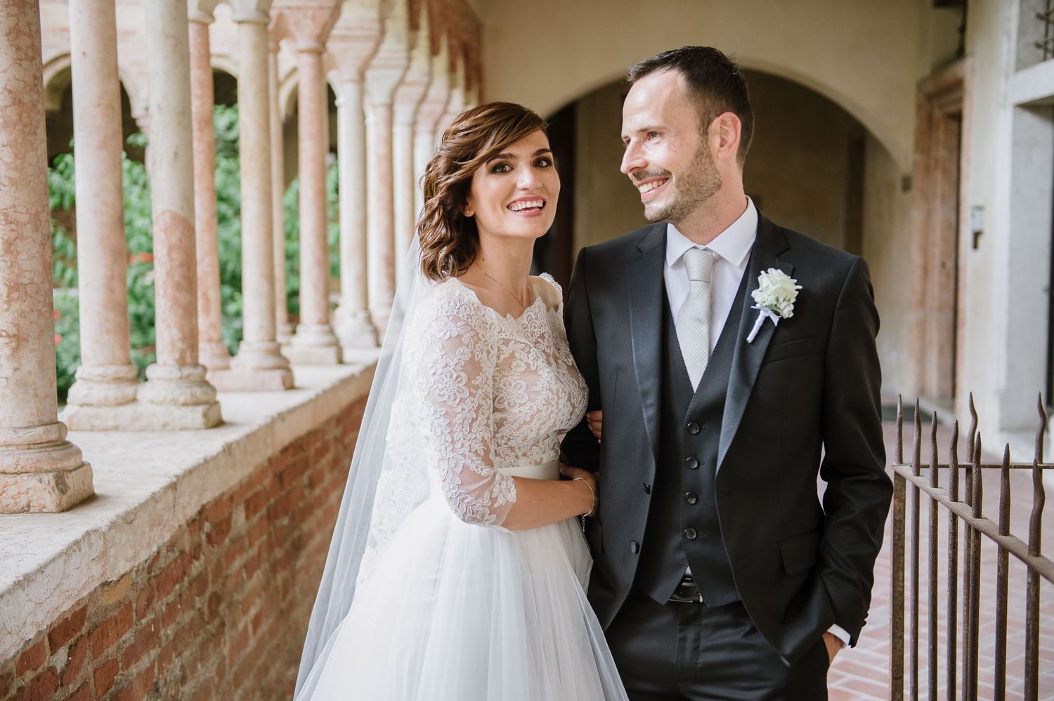 Bride and groom posing together at a classic colonnade venue, bride in lace gown, groom in black suit with boutonniere.