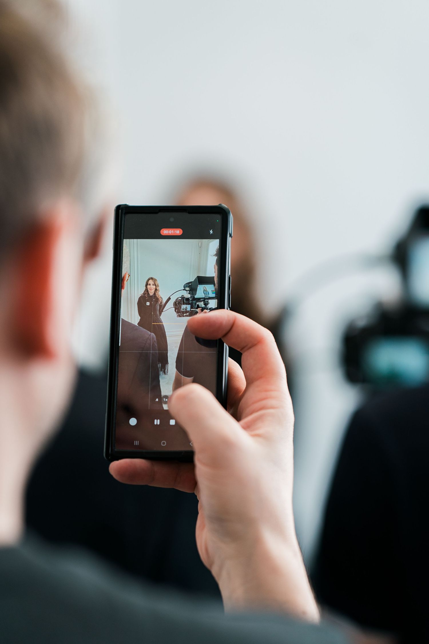 Person filming a woman with a smartphone during a professional video shoot with camera equipment visible.