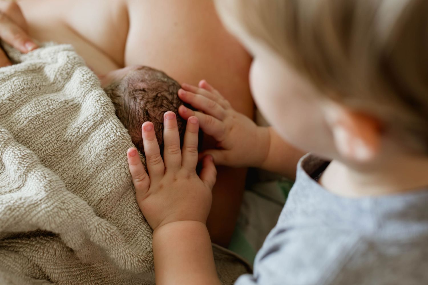 A young child gently touches a newborn baby's head with both hands in a tender moment.