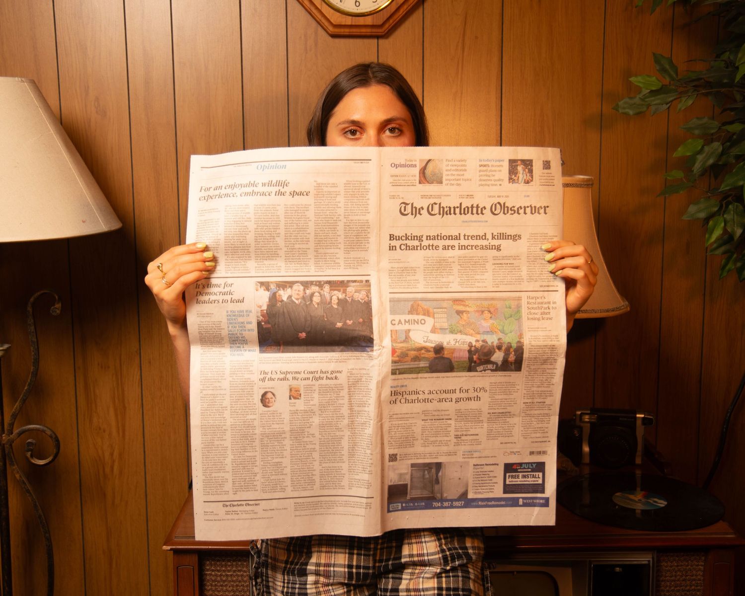 Person holding up The Charlotte Observer newspaper, covering their face, standing in front of wood-paneled wall.