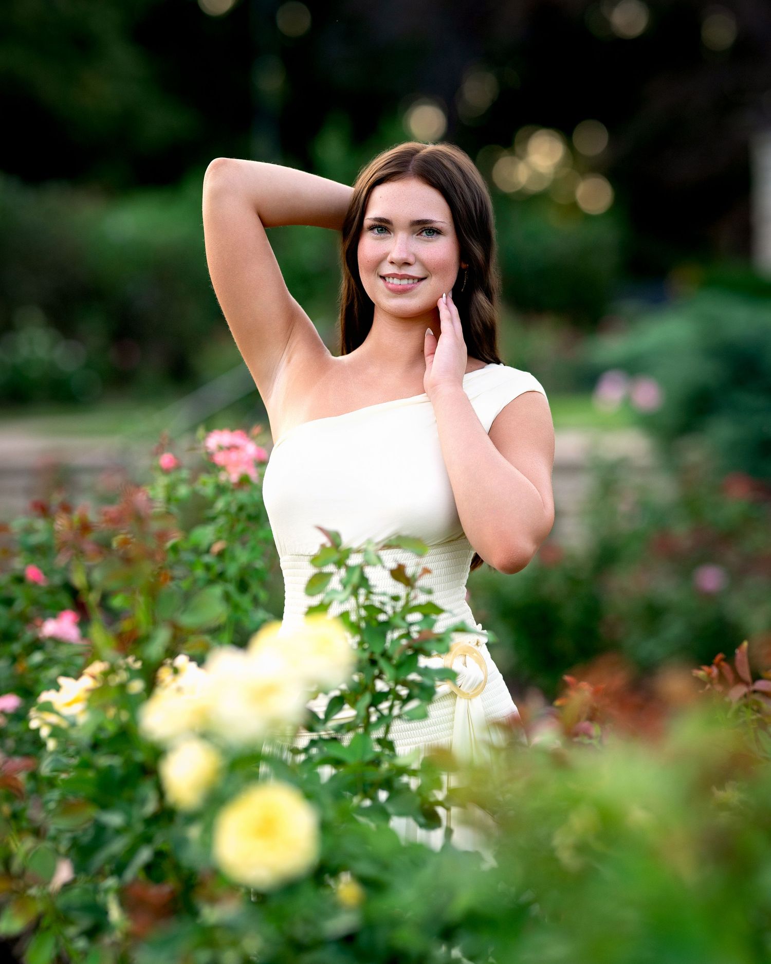 Young woman in white dress posing gracefully among colorful roses in a lush garden setting. Jen Welker, Kansas City Senior Portraits