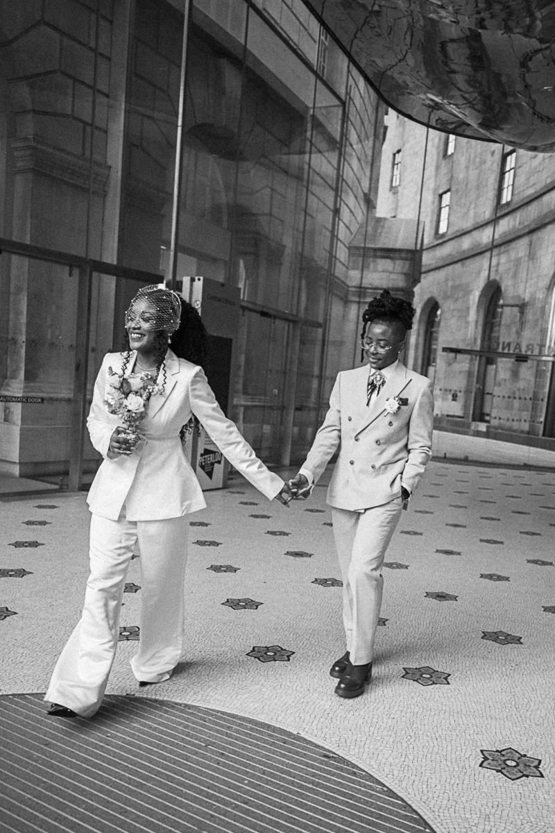 Two brides in white suits hold hands and walk joyfully outside a modern building in a black and white wedding photo.