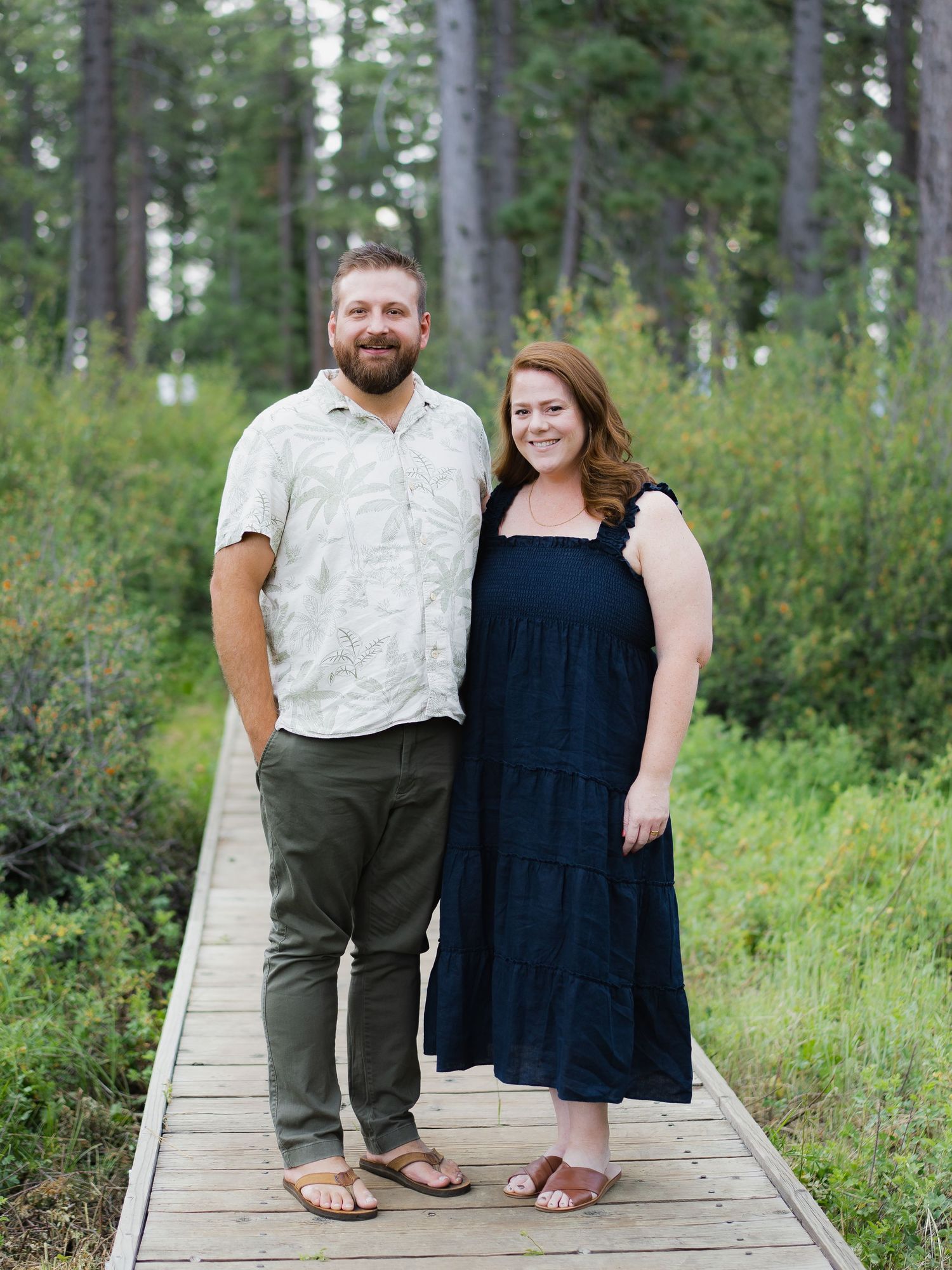 A smiling couple stands on a wooden boardwalk surrounded by lush green forest trees.