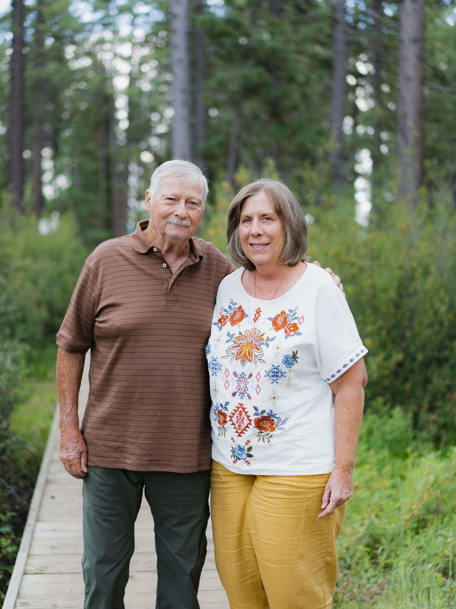 Elderly couple smiling together on a wooden boardwalk path surrounded by tall pine trees in a forest setting.