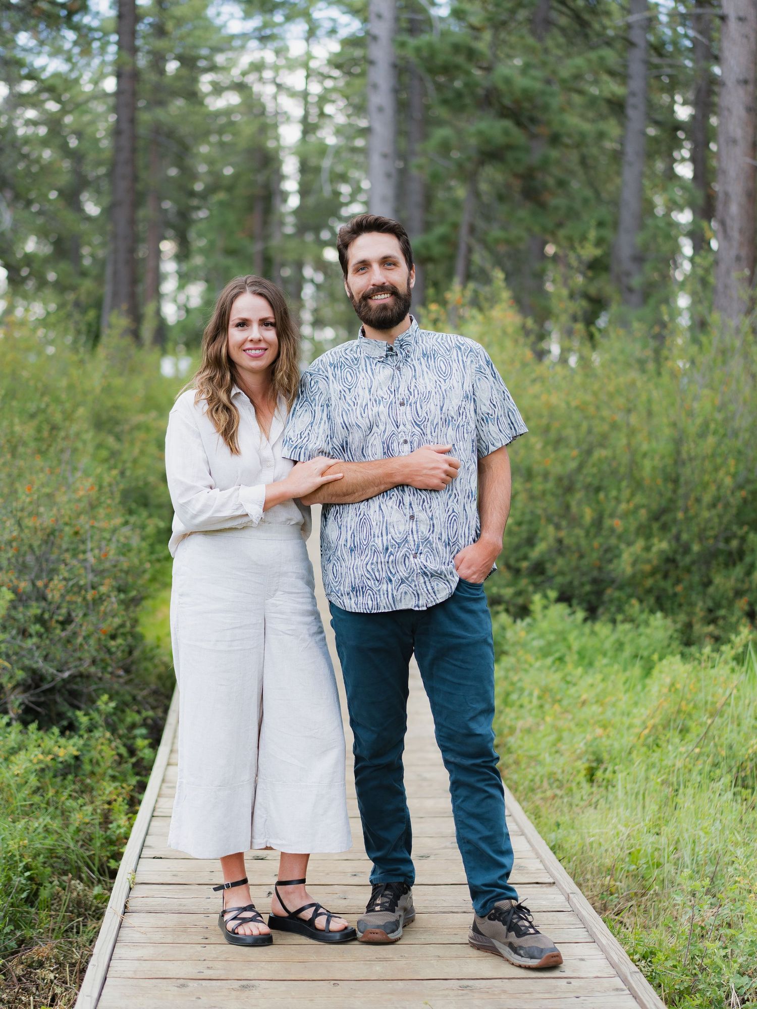 A smiling couple stands on a wooden boardwalk surrounded by lush green forest trees in a casual outdoor portrait.