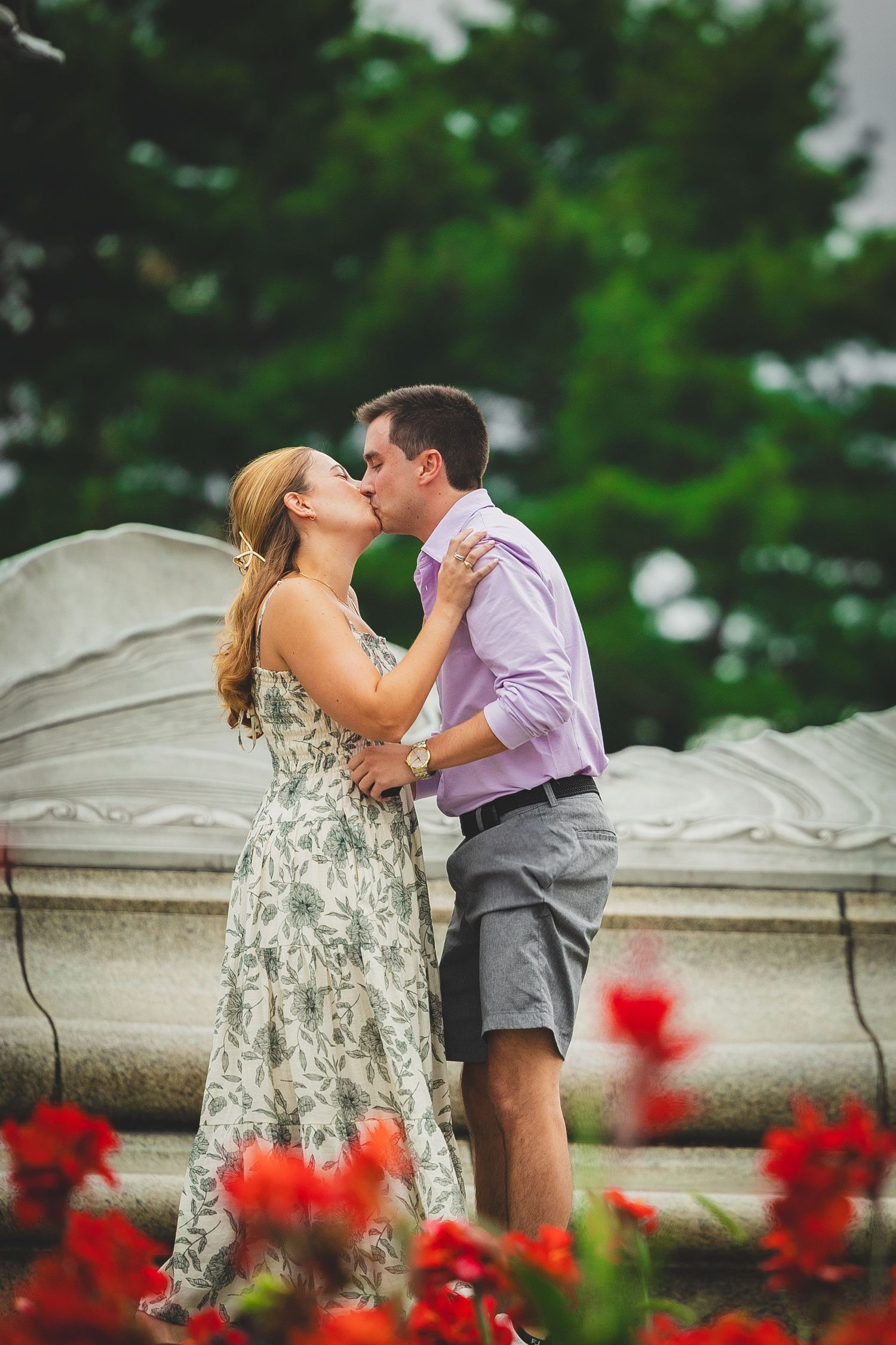 A couple shares a romantic kiss surrounded by red flowers and lush green trees near a stone monument outdoors.