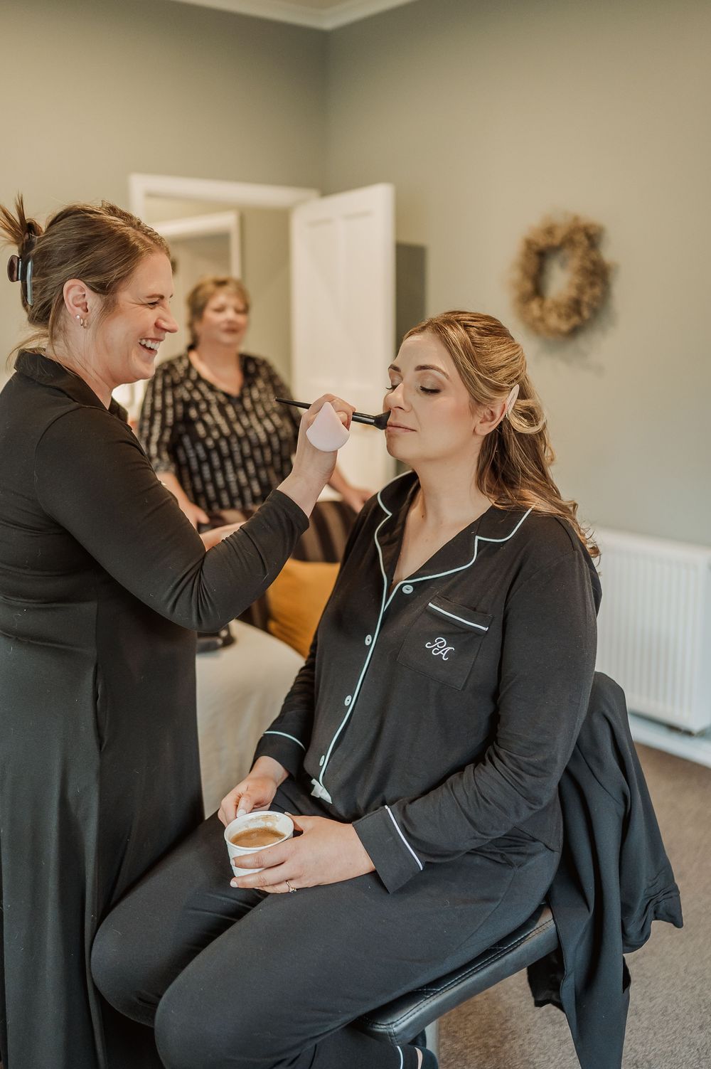 Bride getting makeup applied by artist in black outfit while holding coffee cup in cozy room.