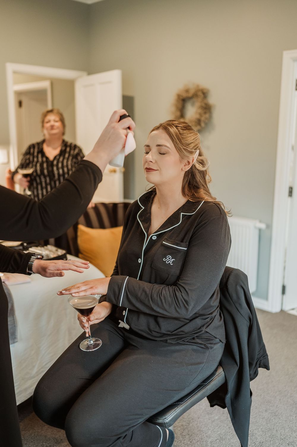 Bride in black monogram pajamas holds wine glass while getting makeup done by artist in elegant room.