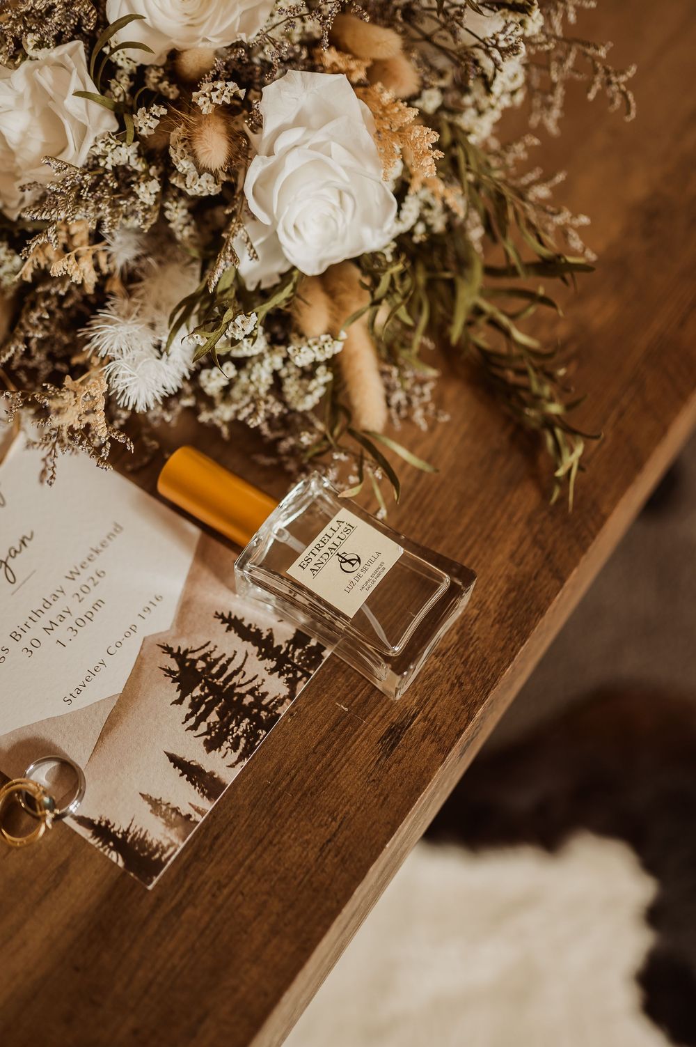 Rustic wedding flat lay with perfume bottle, dried floral bouquet, invitation, and gold ring on wooden table.