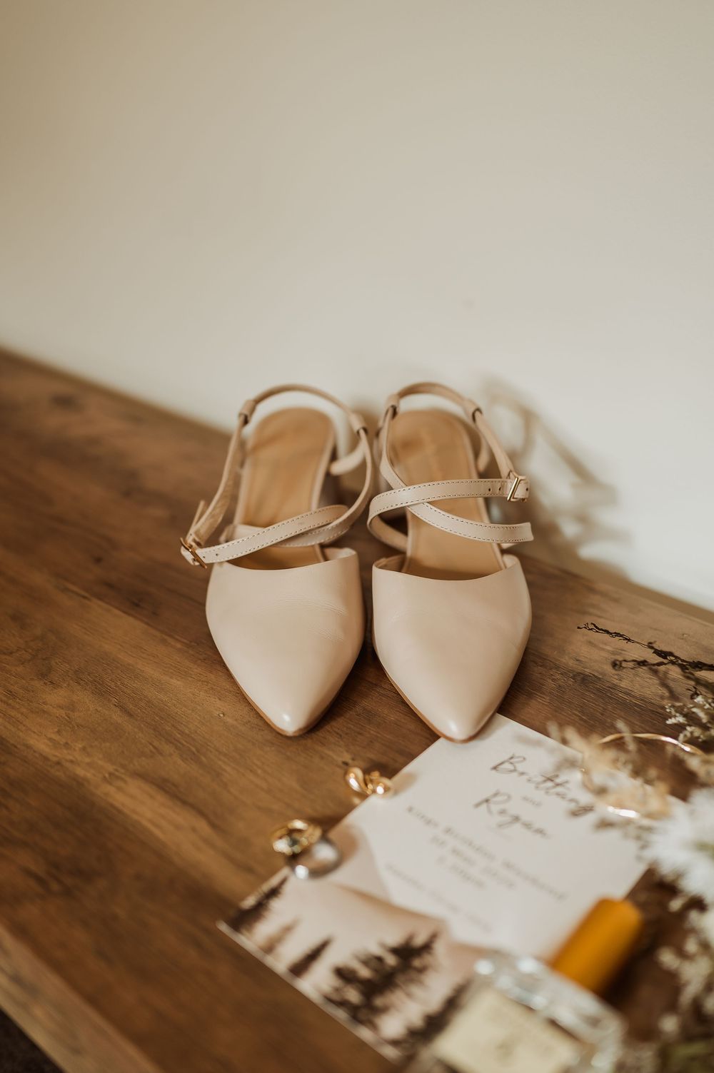 Nude pointed-toe slingback flats with cross straps on a wooden table beside wedding invitation cards.