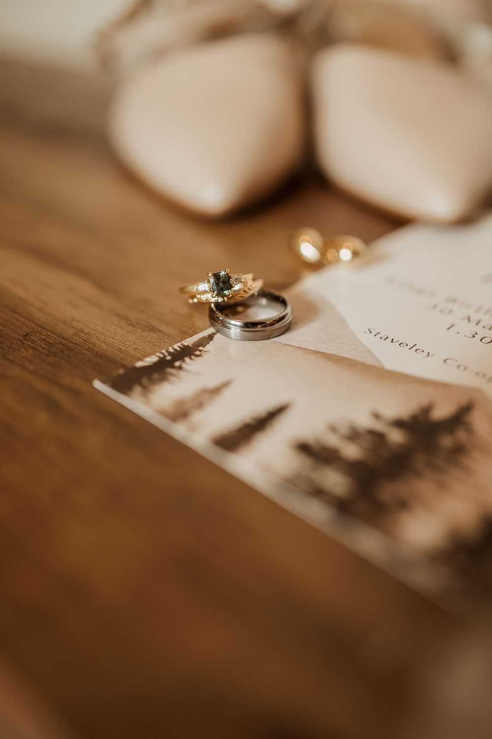 Close-up of wedding rings and earrings resting on an invitation card with nude bridal heels in the background.