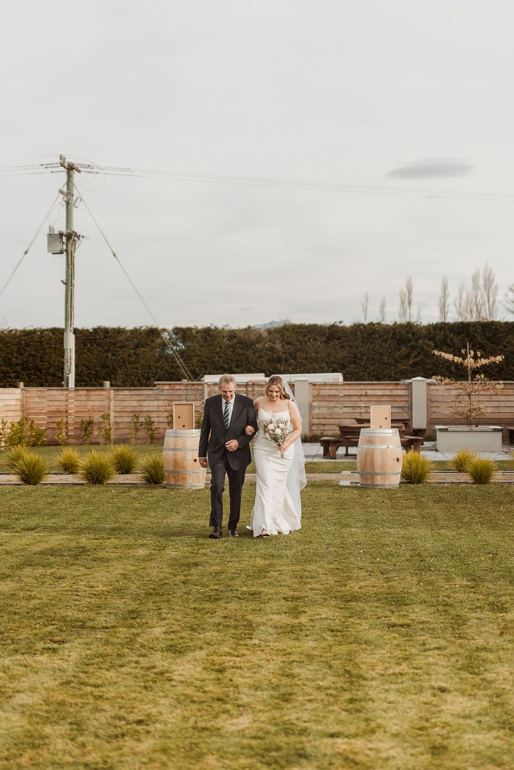 Bride and groom walking across green lawn at outdoor wedding venue with wooden barrels and hedge backdrop.
