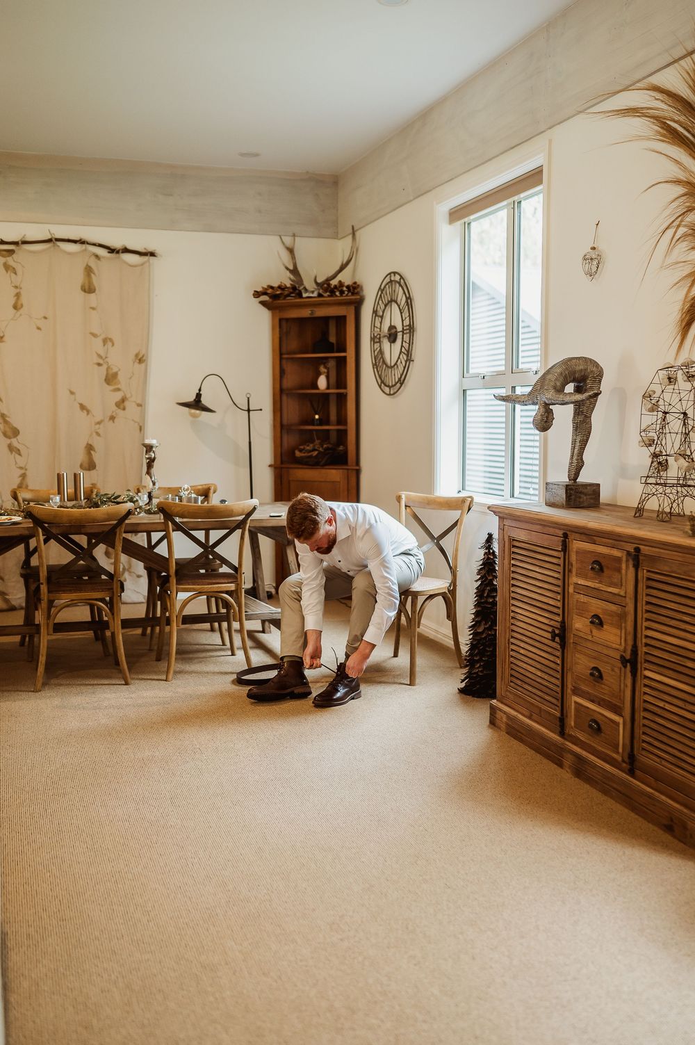 Groom in white shirt ties dress shoes in rustic wooden dining room before wedding ceremony.
